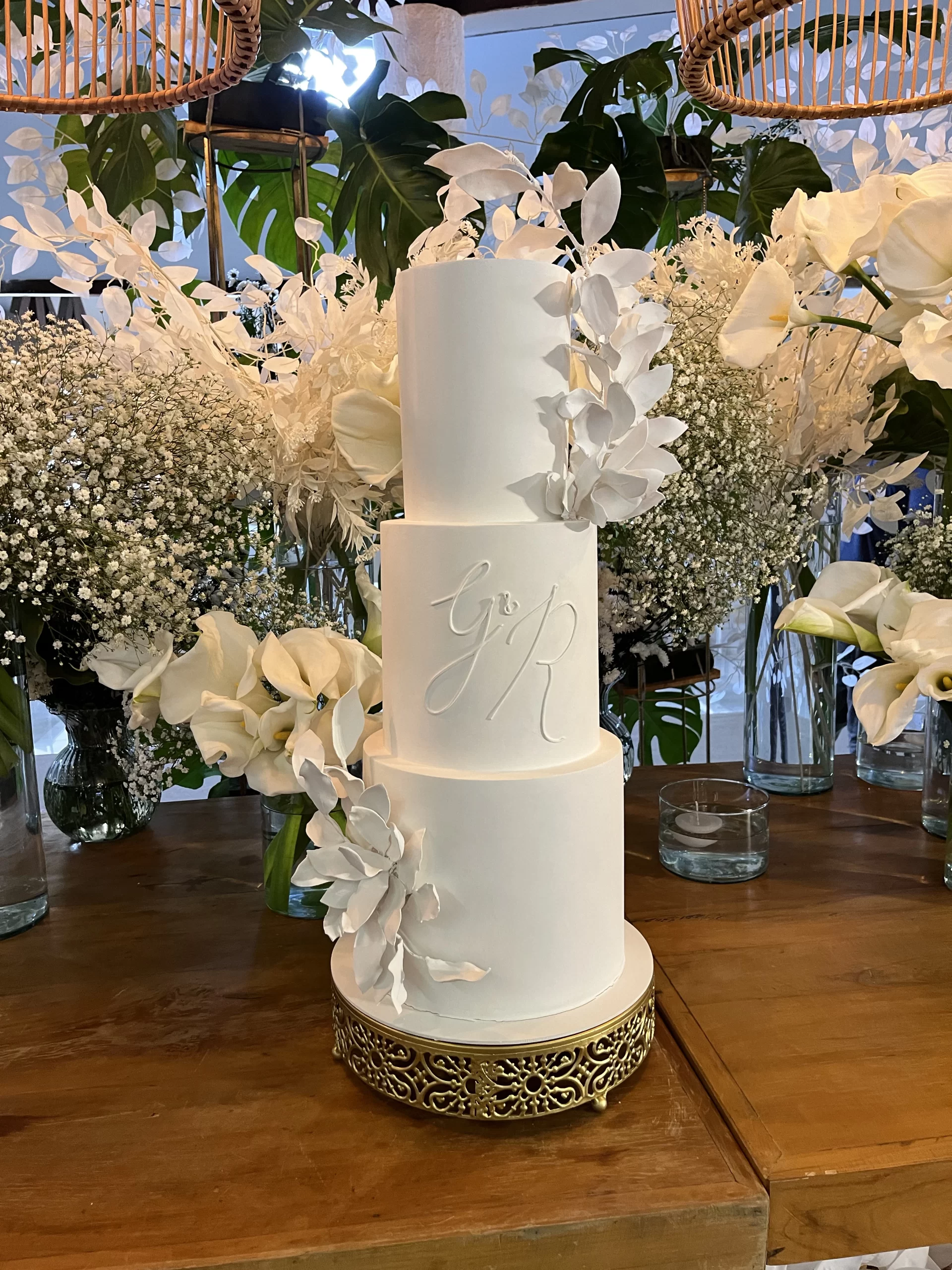 Bolo de casamento branco minimalista com iniciais e folhagens brancas.
